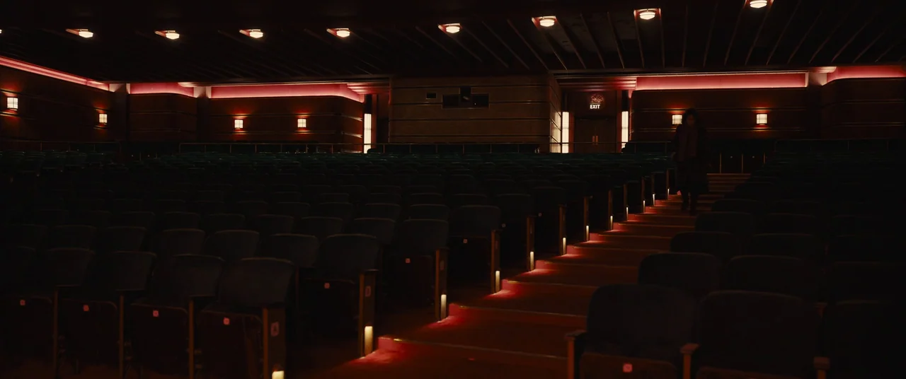 Empire of Light (2022) movie still: wide — A dark movie theater auditorium features rows of empty seats and red carpeted aisles. A lone woman walks…