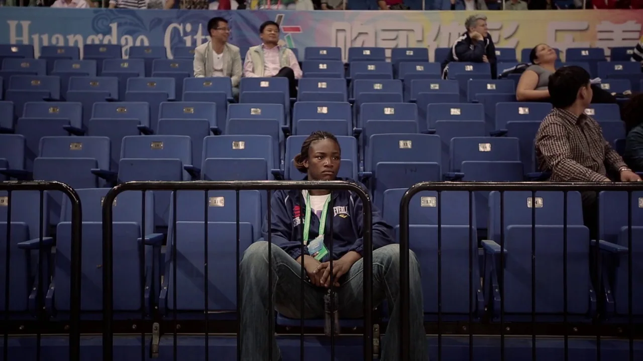 T-Rex (2015) movie still: medium — A rows of blue stadium seats feature an athletic woman sitting near the front. Other spectators sit scattered in…
