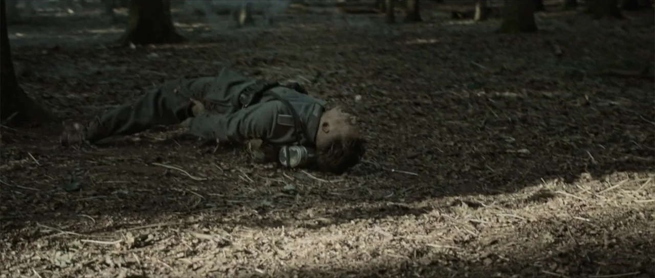 Sin retorno (2009) movie still: fullbody — A dense forest floor covered in dead leaves and tree roots under dark trees. A soldier in uniform lies…