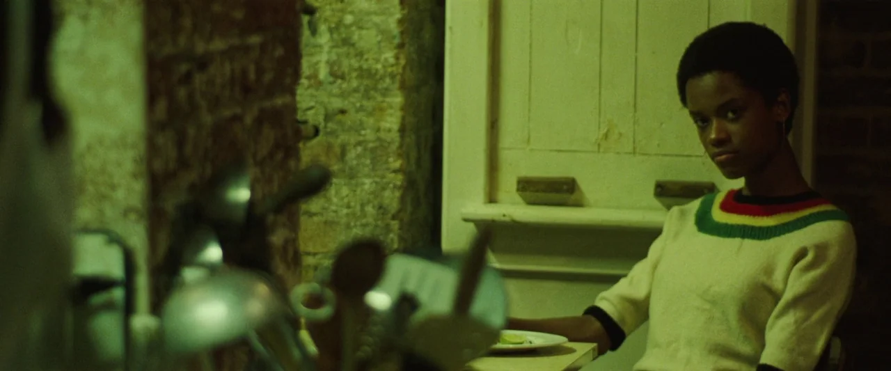 Mangrove (2020) movie still: medium — A dimly lit room features a rough brick wall and a white door frame. A young black woman sits at a table while…