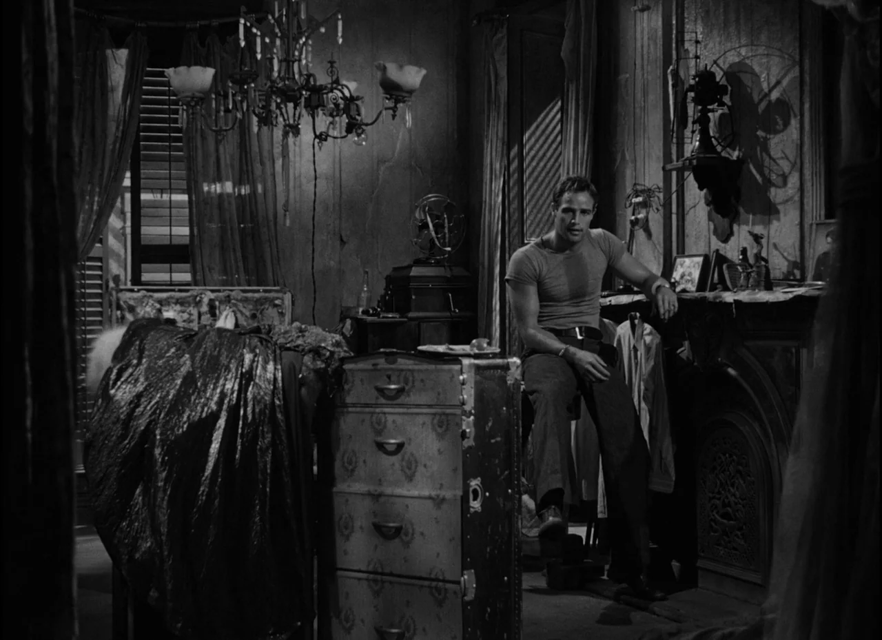A Streetcar Named Desire (1951) movie still: fullbody — A dimly lit room contains a cluttered dresser, a ceiling chandelier, and a metal fan. A…