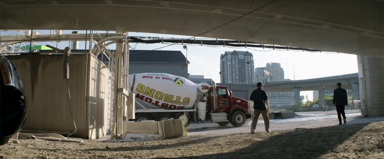 The Equalizer (2014) movie still: wide — A gravel-covered area sits beneath a heavy concrete highway overpass with city buildings visible. Two men…