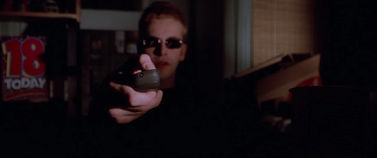 Hackers (1995) movie still: medium — A dimly lit room with an '18' birthday card visible on a shelf. A young man sits in the dark while pointing a…