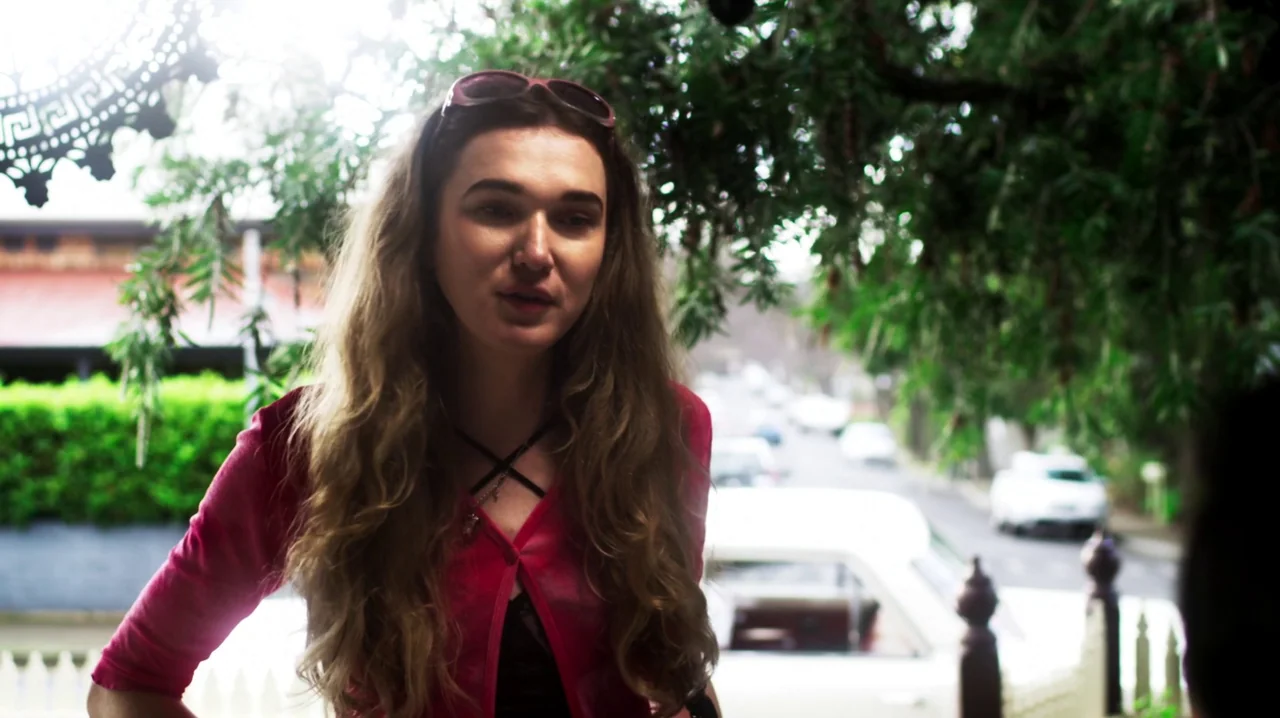 Carnage for Christmas (2024) movie still: medium — A woman stands on a porch with greenery and a blurred street behind her. The woman is speaking…
