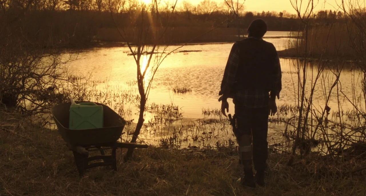 The Alchemist Cookbook (2016) movie still: fullbody — A man stands on the grassy edge of a marshy lake during sunset. A metal wheelbarrow containing…