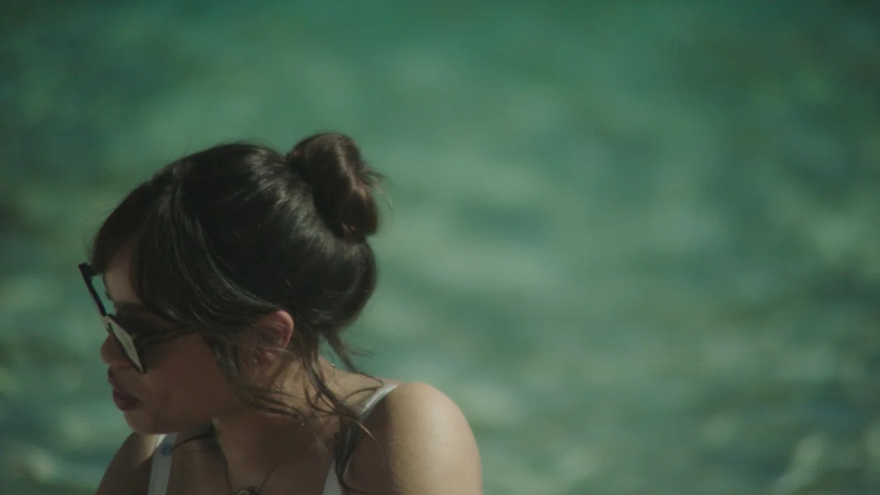 Infinity Pool (2023) movie still: closeup — A woman stands submerged in clear, shallow turquoise water. She wears sunglasses while looking toward…