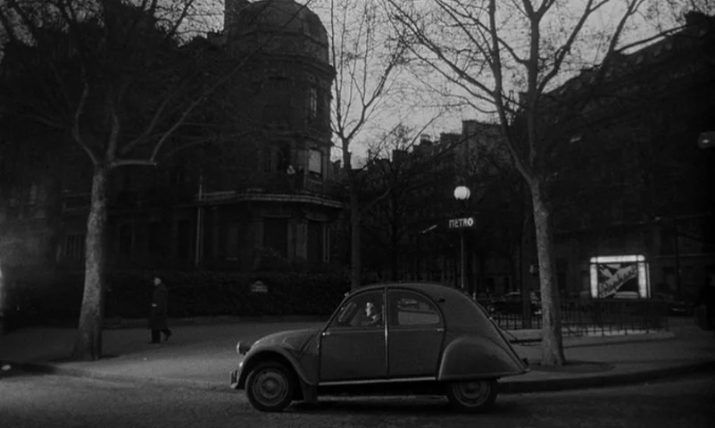 Eyes Without A Face (1960) movie still: wide — A Parisian city square features bare trees, historic stone buildings, and a metro entrance. A man…