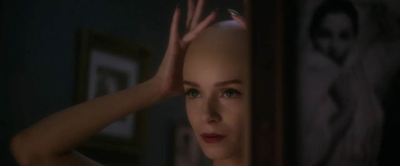 Starry Eyes (2014) movie still: closeup — A dimly lit room with a mirror reflecting a woman's face. The subject is a bald woman holding her head…