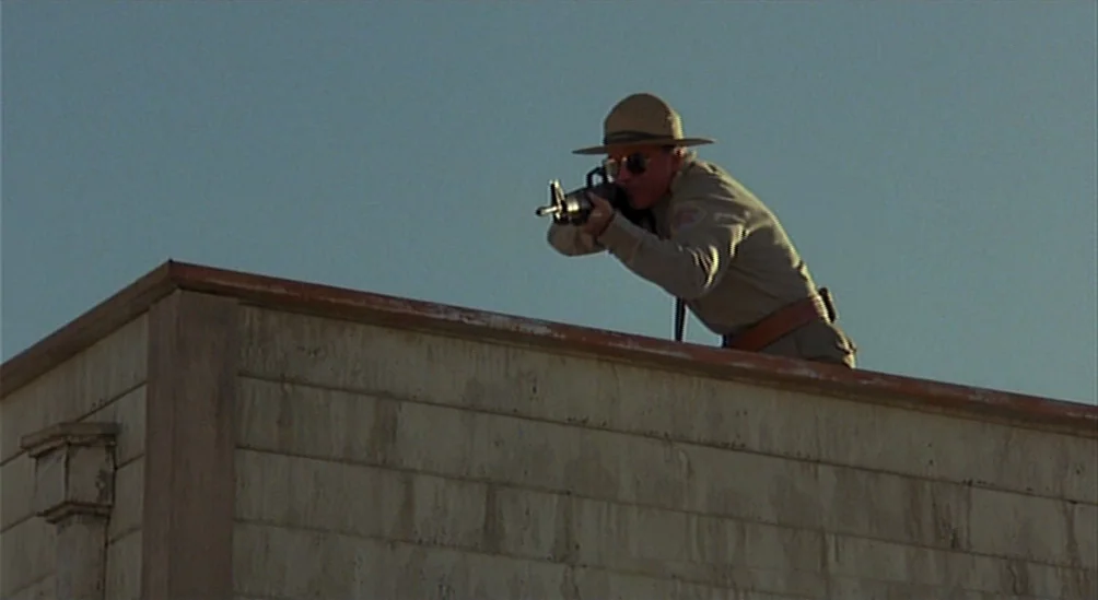 Near Dark (1987) movie still: medium — A man in a tan uniform kneels at the edge of a weathered building. He aims an assault rifle toward the…