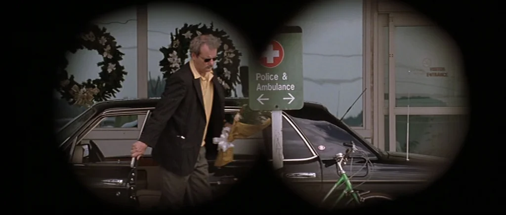 Rushmore (1998) movie still: medium — A man exits a car in a parking lot near a building entrance with wreaths. He is holding a bouquet and walking…