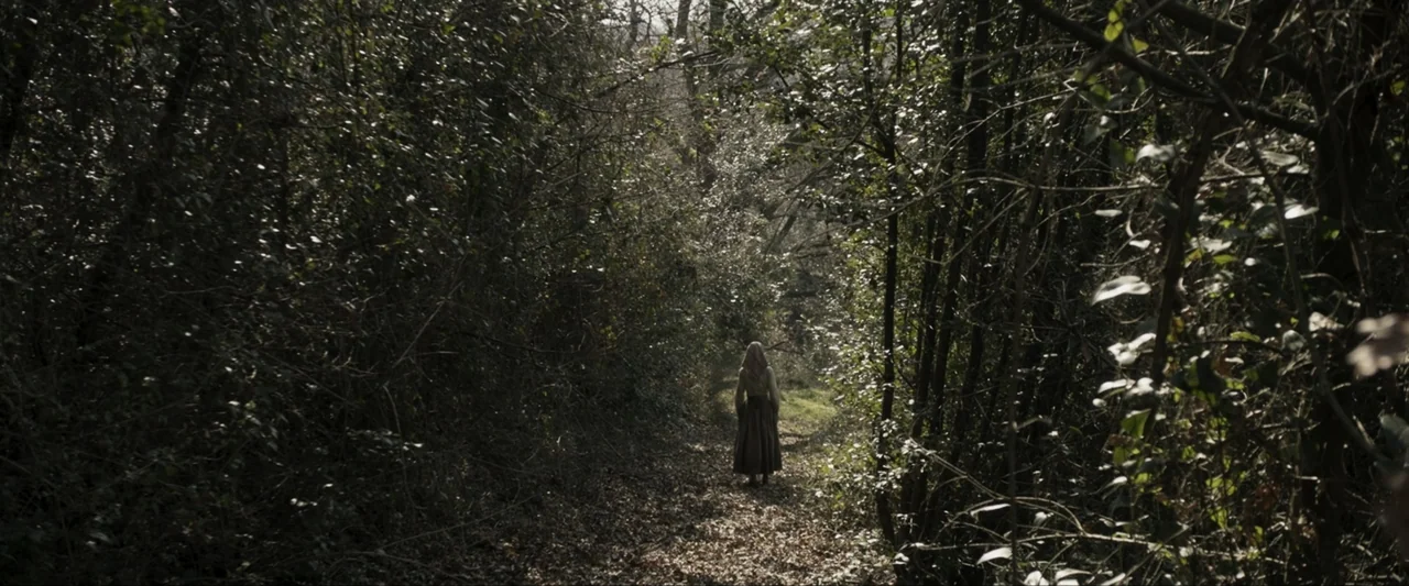 Fatima (2020) movie still: wide — A narrow dirt path winds through a dense thicket of trees and overgrown bushes. A woman in period clothing walks…