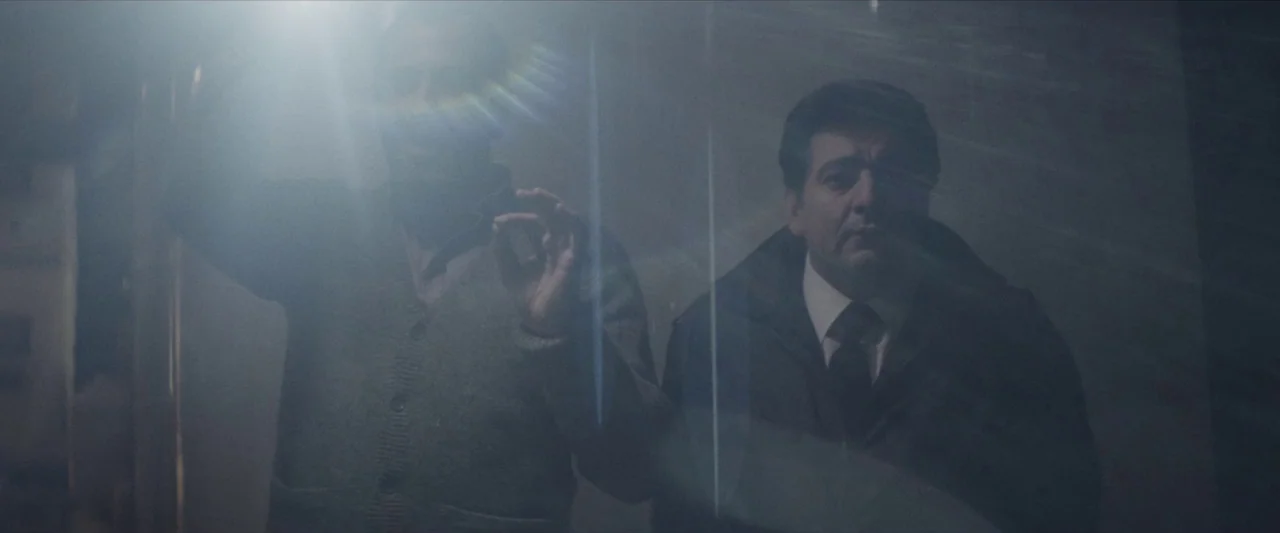 Chevalier (2015) movie still: medium — A dimly lit lobby area viewed through a glass partition with light reflections. Two men stand together inside…