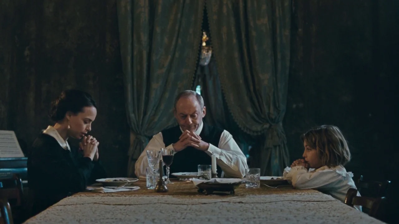 Childhood of a Leader (2015) movie still: medium — A dimly lit room with heavy green fabric curtains and a dining table. A woman, an older man, and…