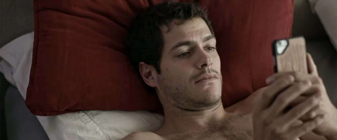 Do It Like An Hombre (2017) movie still: closeup — A man lies in a bed with a dark red pillow behind his head. The man looks at his smartphone…