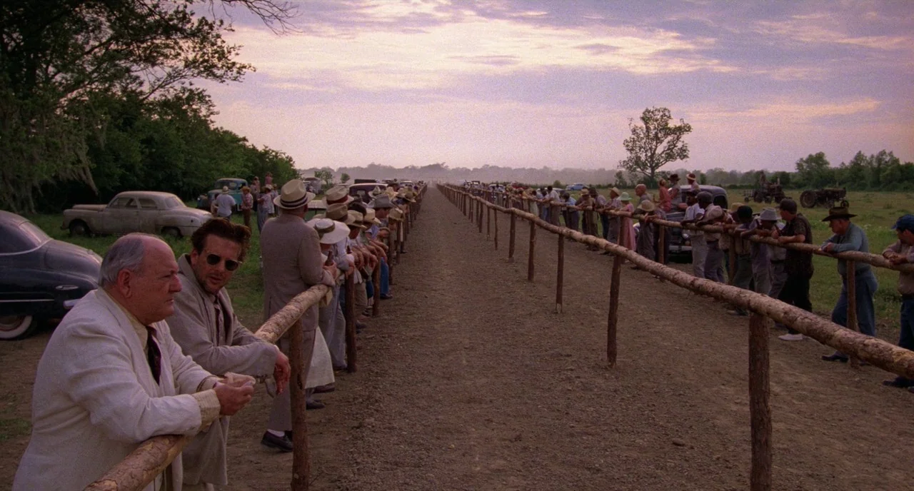 Angel Heart (1987) movie still: wide — A long, straight wooden fence runs through an open field at golden hour. A large crowd of people in…
