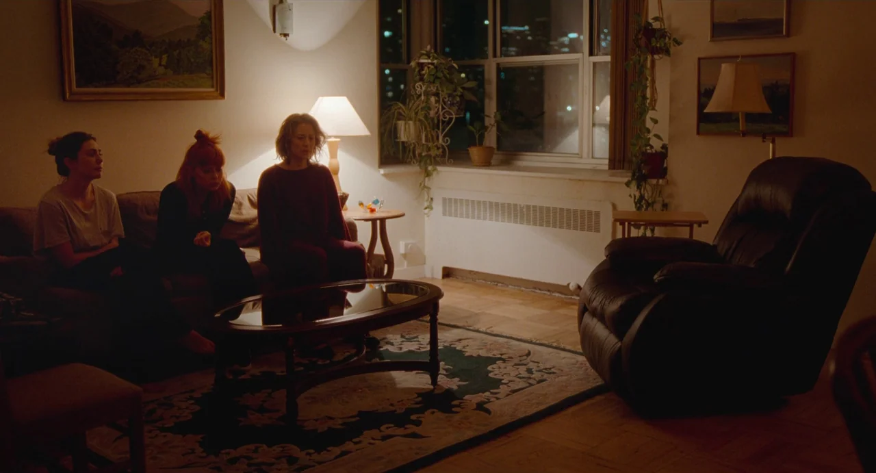 His Three Daughters (2023) movie still: wide — An apartment interior featuring a couch with three seated women facing an empty recliner. Potted…