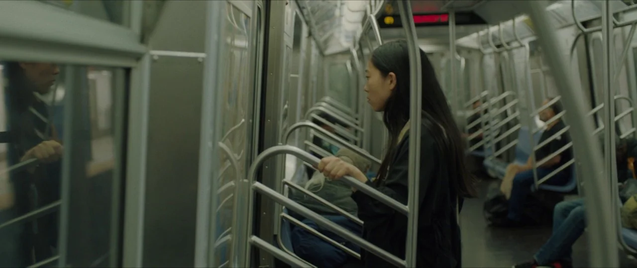 The Farewell (2019) movie still: medium — The interior of a New York City subway car with metal handrails and seats. A young woman stands in the…