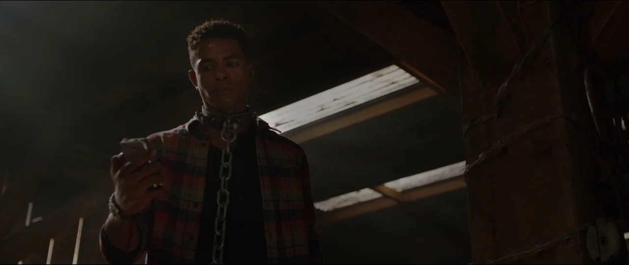 Jigsaw (2017) movie still: medium — A dark wooden structure with horizontal light slats visible through the roof. A young man stands in the center…