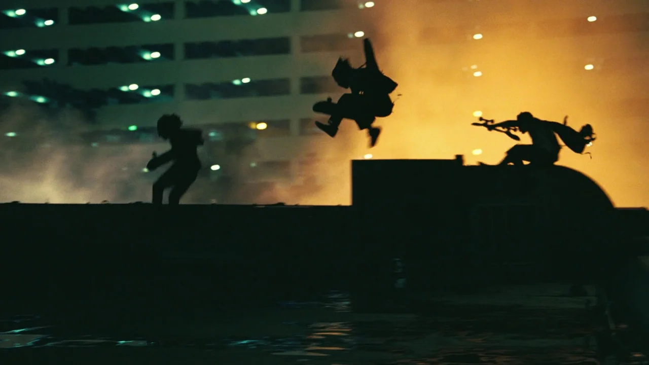 One Battle After Another (2025) movie still: wide — A dark rooftop parking level features a warm glowing orange background with building lights.…