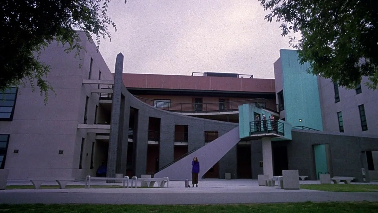 Secretary movie still: wide — A modern academic building with a distinctive curved grey wall and teal structural elements. A solitary woman stands…