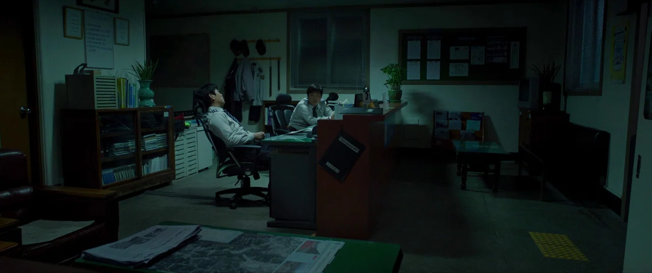The Wailing (2016) movie still: wide — A dimly lit security station filled with wooden furniture and office documents at night. Two security guards…