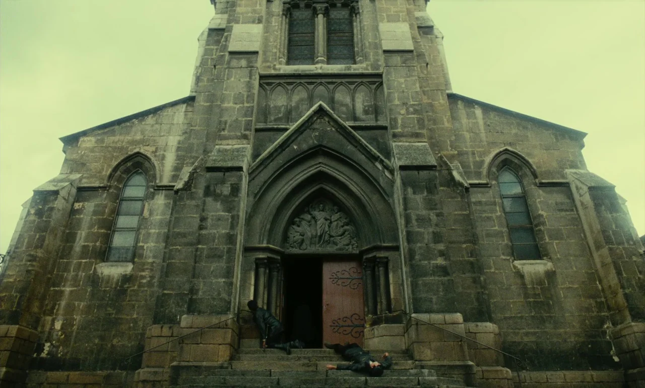 Litan (1982) movie still: wide — A stone gothic church exterior features weathered masonry and a central arched wooden doorway. Two individuals lie…