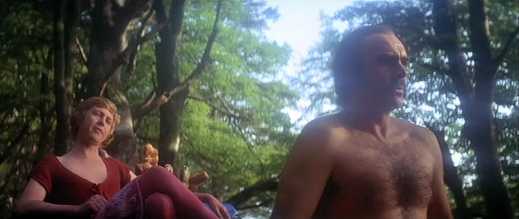 Zardoz (1974) movie still: medium — A sunlit woodland area filled with dense trees and green foliage. A bare-chested man and a person in red…