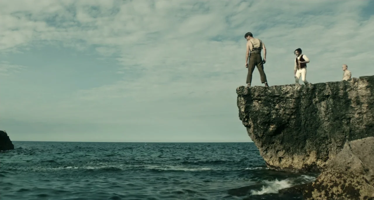 Pinocchio (2022) movie still: wide — A high rocky cliff face overlooks a calm expanse of open ocean water. Three men in period-appropriate attire…