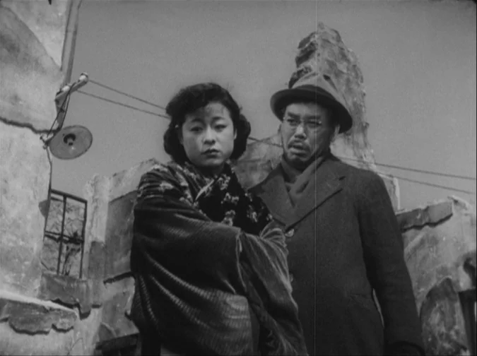 Drunken Angel (1948) movie still: medium — A ruined environment with crumbling concrete walls and debris under an open sky. A woman and a man stand…
