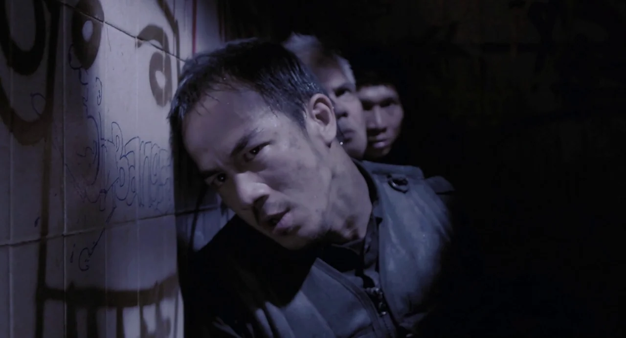 The Raid (2012) movie still: medium — A dark subterranean space features tiled walls covered in black graffiti. Three men huddle together in the dim…