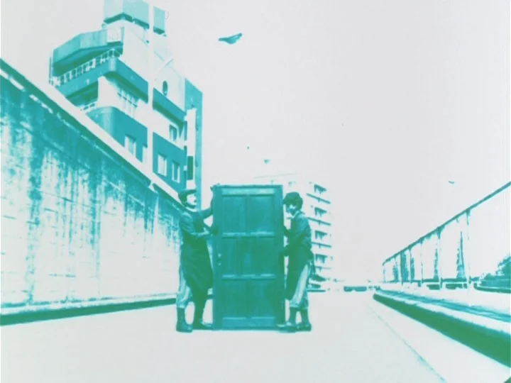Labyrinth Tale (1975) movie still: wide — A wide shot shows a quiet asphalt street flanked by concrete walls and buildings. Two figures in matching…