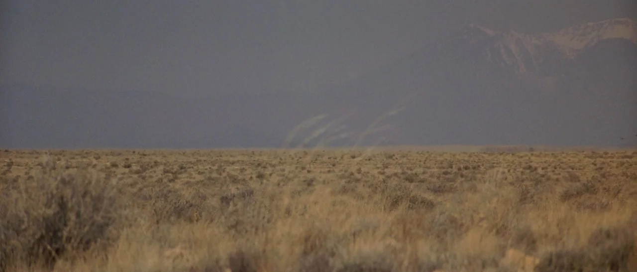 Starman (1984) movie still: establishing — A vast, flat stretch of high-desert scrubland extends toward distant, hazy mountain peaks. Dry grass and…