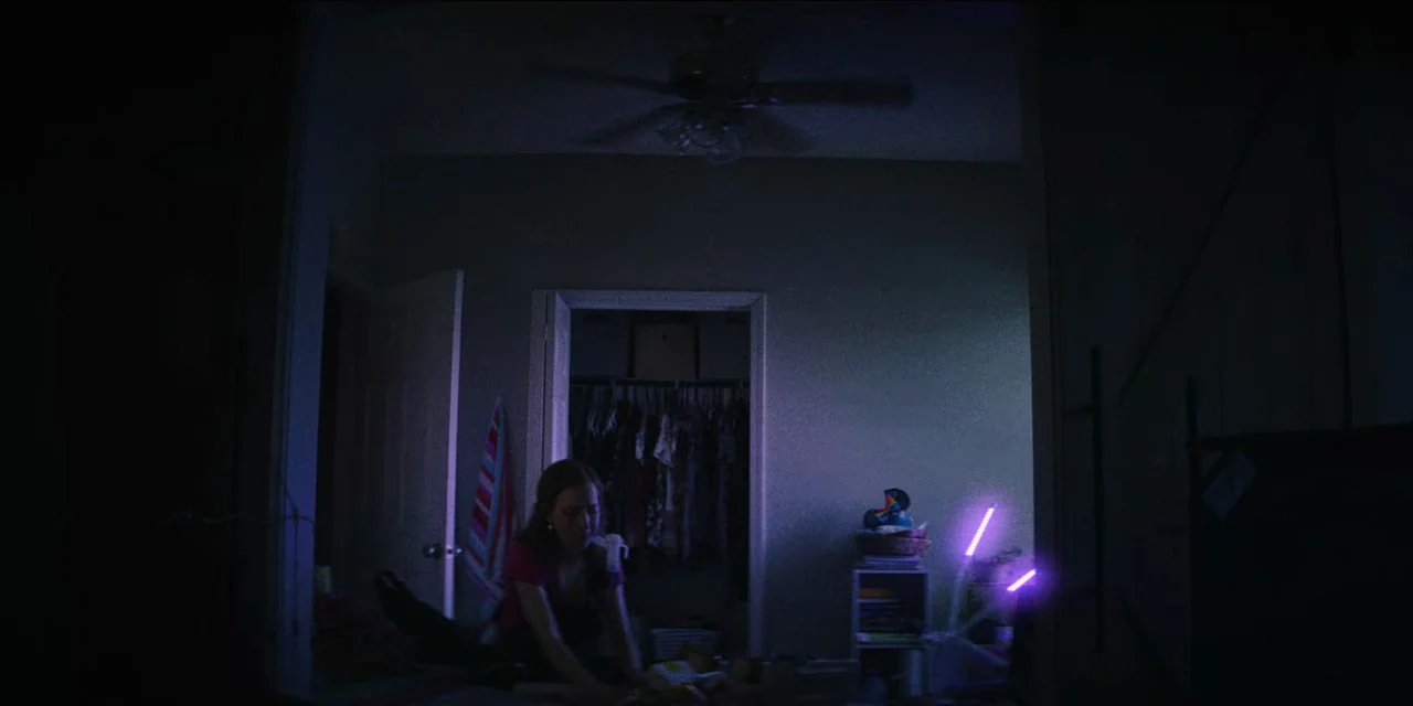 Spermworld (2024) movie still: wide — A dimly lit bedroom features an open closet and a ceiling fan overhead. A young woman sits on the floor…