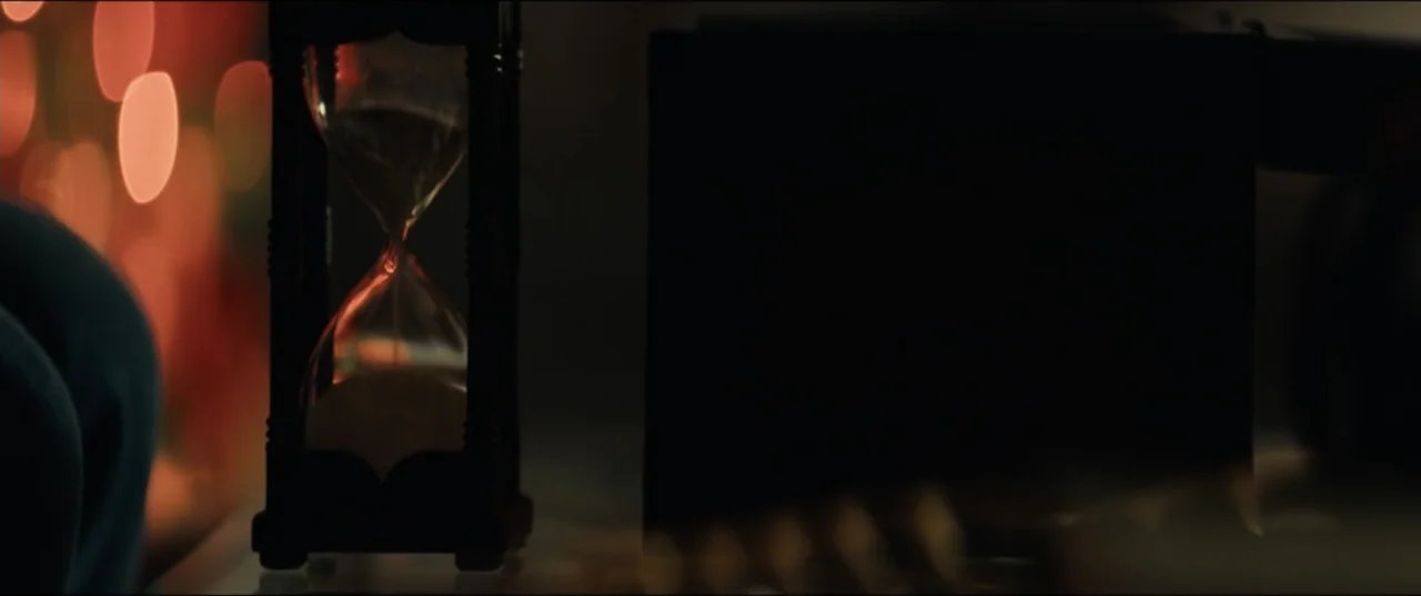 Vicious (2025) movie still: closeup — A dark indoor space contains a wooden hourglass sitting on a reflective surface. The sand flows through the…