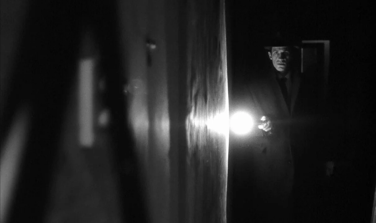 Le Doulos (1962) movie still: medium — A dark, narrow interior space features a solid wall occupying the foreground. A man in a hat holds a…