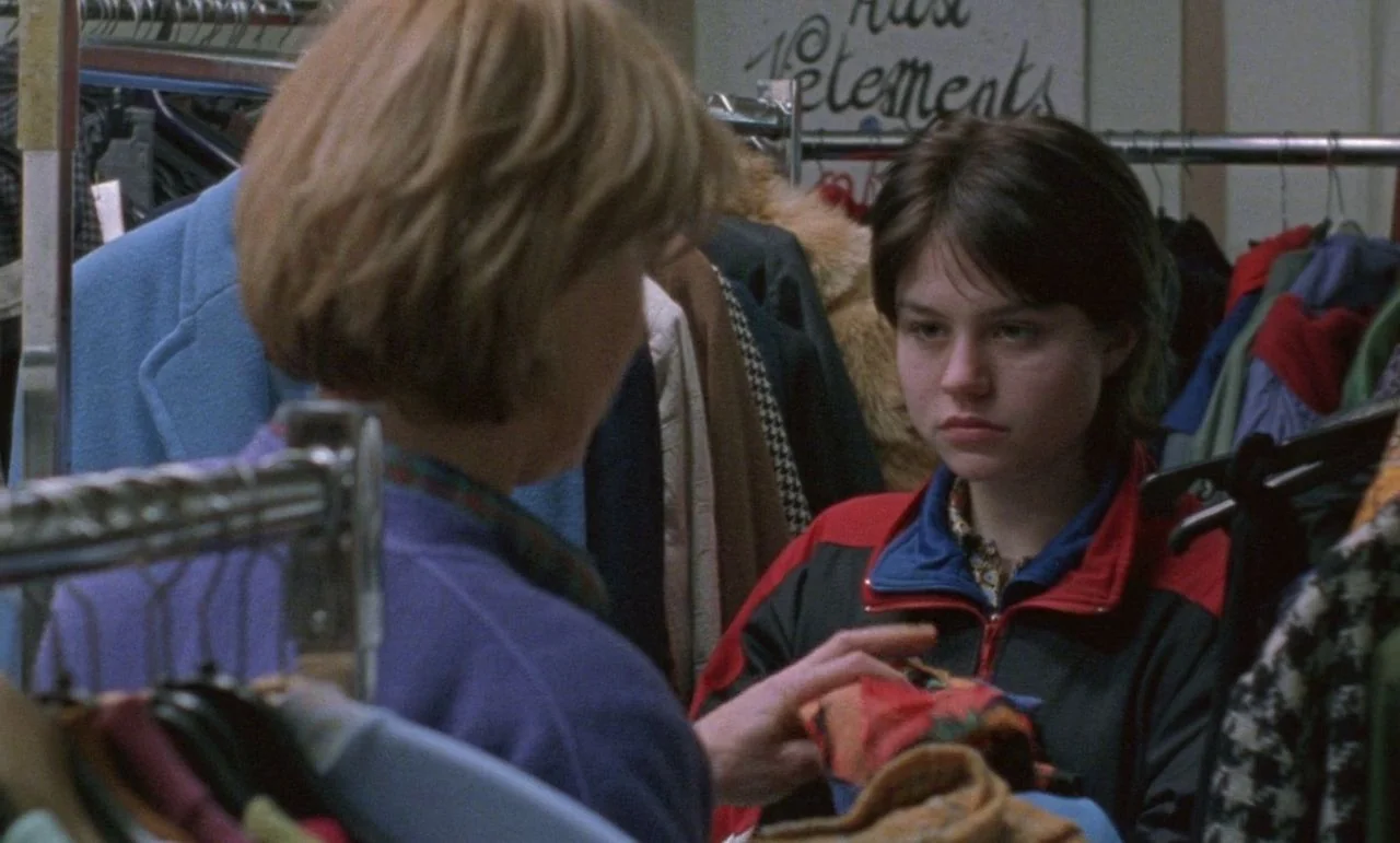 Rosetta (1999) movie still: medium — A cluttered second-hand clothing store features racks packed with garments and handwritten paper signage. A…