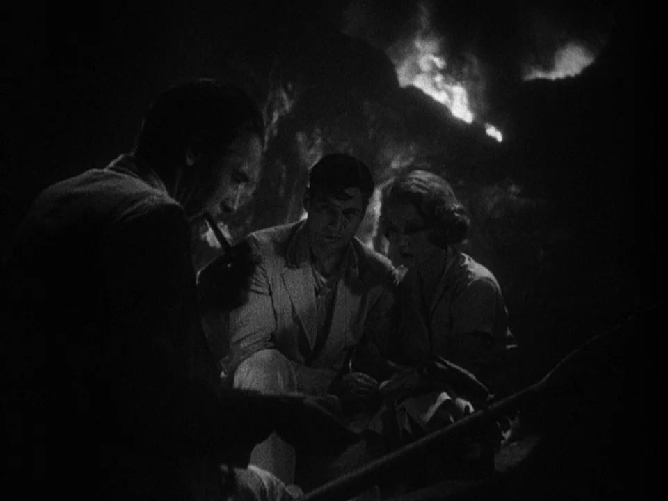 Island of Lost Souls (1932) movie still: medium — Three individuals are clustered in a dimly lit, jagged rocky space. A fire burns behind them in…