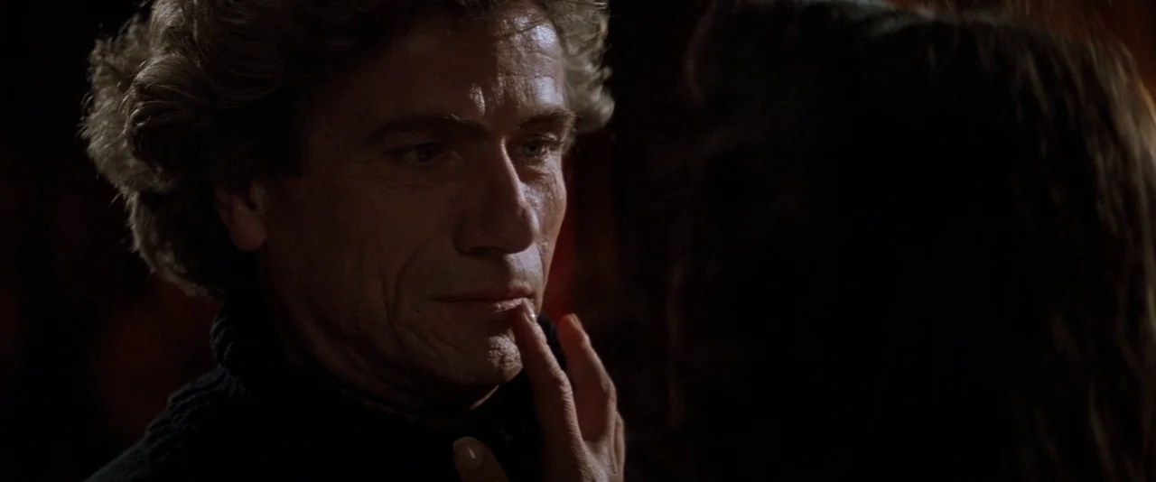 In The Mouth of Madness (1994) movie still: closeup — A dimly lit interior space with dark, indistinct surroundings. A woman's hand reaches out to…