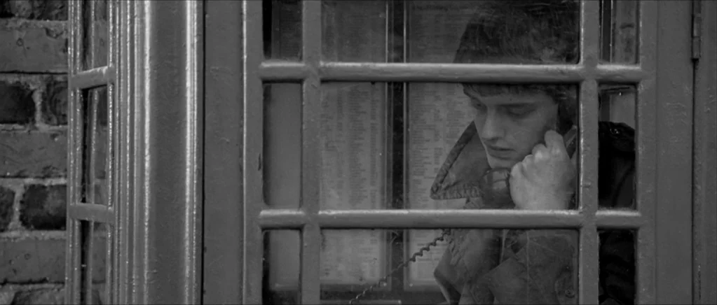 Control movie still: medium — A man stands inside a classic red British style public telephone box. He speaks into the handset while framed by the…