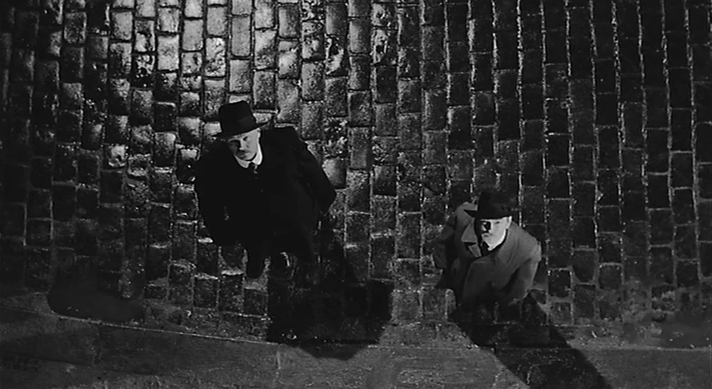 Kafka (1991) movie still: fullbody — A wet cobblestone street at night is illuminated by a sharp overhead light source. Two men in hats and coats…