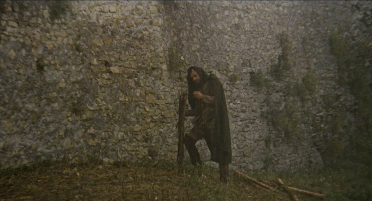 Beatrice Cenci (1969) movie still: fullbody — A man stands before a tall, weathered stone wall with small patches of moss. The person holds a stack…