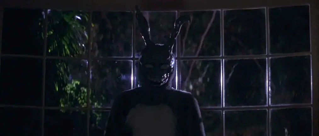 Donnie Darko movie still: medium — A dark interior view of a paneled window looking out into the night. A figure wearing a grotesque rabbit mask…