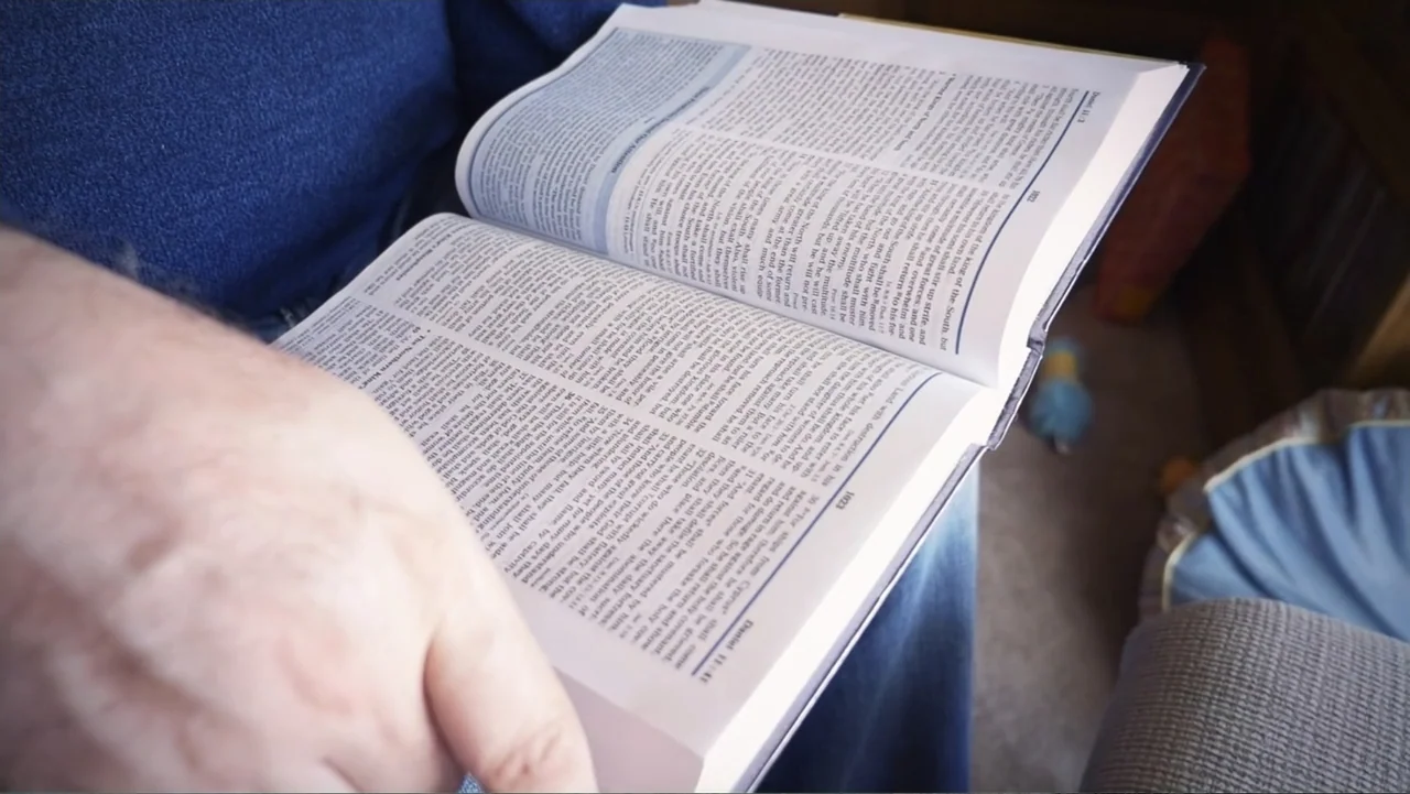 Beware of Angels (2016) movie still: closeup — A person holds an open Bible while sitting in a fabric-covered chair. The pages feature two columns…