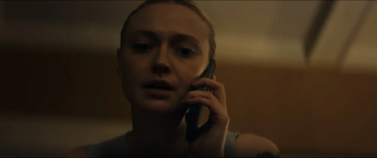 Vicious (2025) movie still: closeup — A dimly lit indoor space with blurred background surfaces. A woman in a sleeveless top holds a smartphone to…