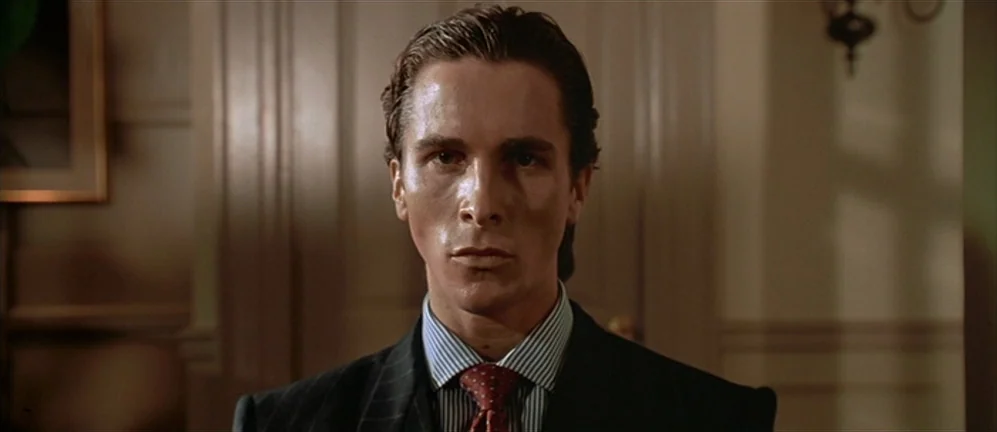 American Psycho (2000) movie still: closeup — A dimly lit indoor hallway with plain wall molding provides a simple backdrop. A man in a suit looks…