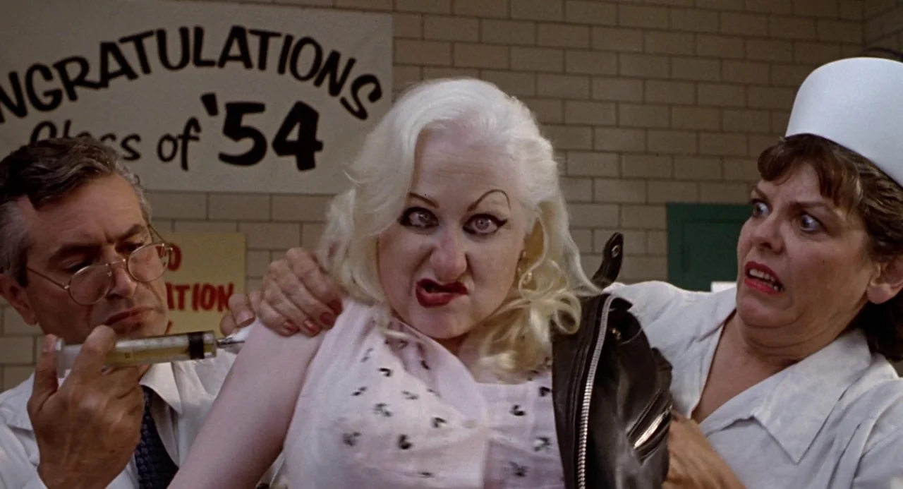 Cry Baby (1990) movie still: medium — A school infirmary with a Congratulations Class of 54 banner hung on the wall. A man administers an injection…