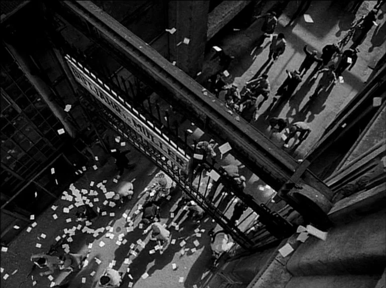 My 20th Century (1989) movie still: establishing — A high-angle view captures a crowd of people moving through an industrial factory gate. Scraps of…