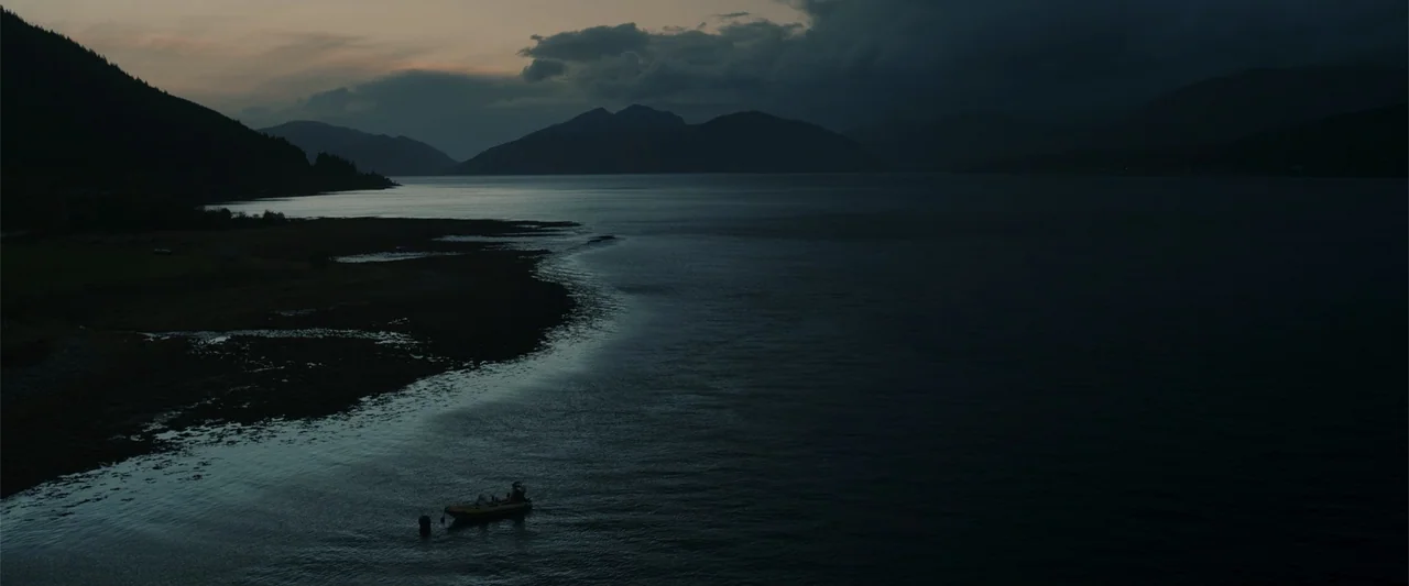 My Son (2021) movie still: establishing — A dark mountainous landscape surrounds a wide expanse of still water at dusk. A small yellow boat sits in…