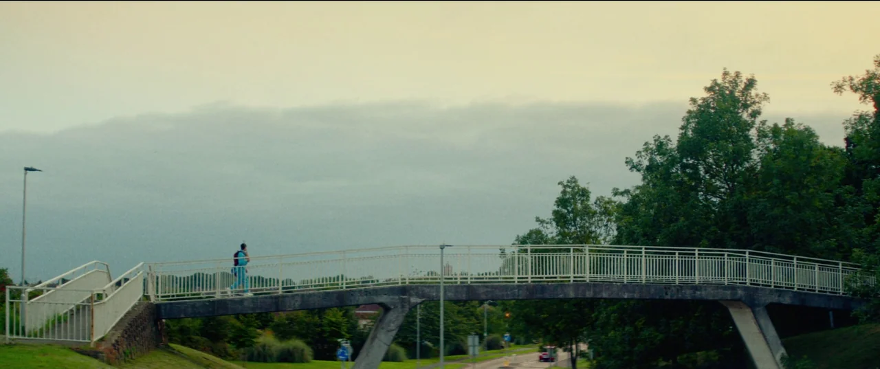 Scrapper (2023) movie still: wide — A concrete footbridge spans over a road, flanked by green trees under cloudy skies. A lone person in a blue…