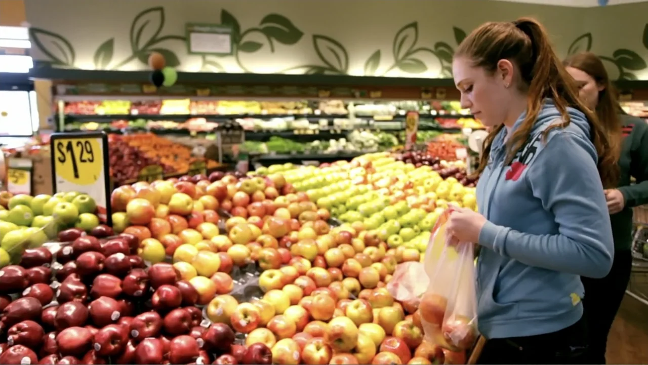 The (Dead Mothers) Club (2014) movie still: medium — A well-lit grocery store aisle features stacked rows of red, yellow, and green apples. A young…